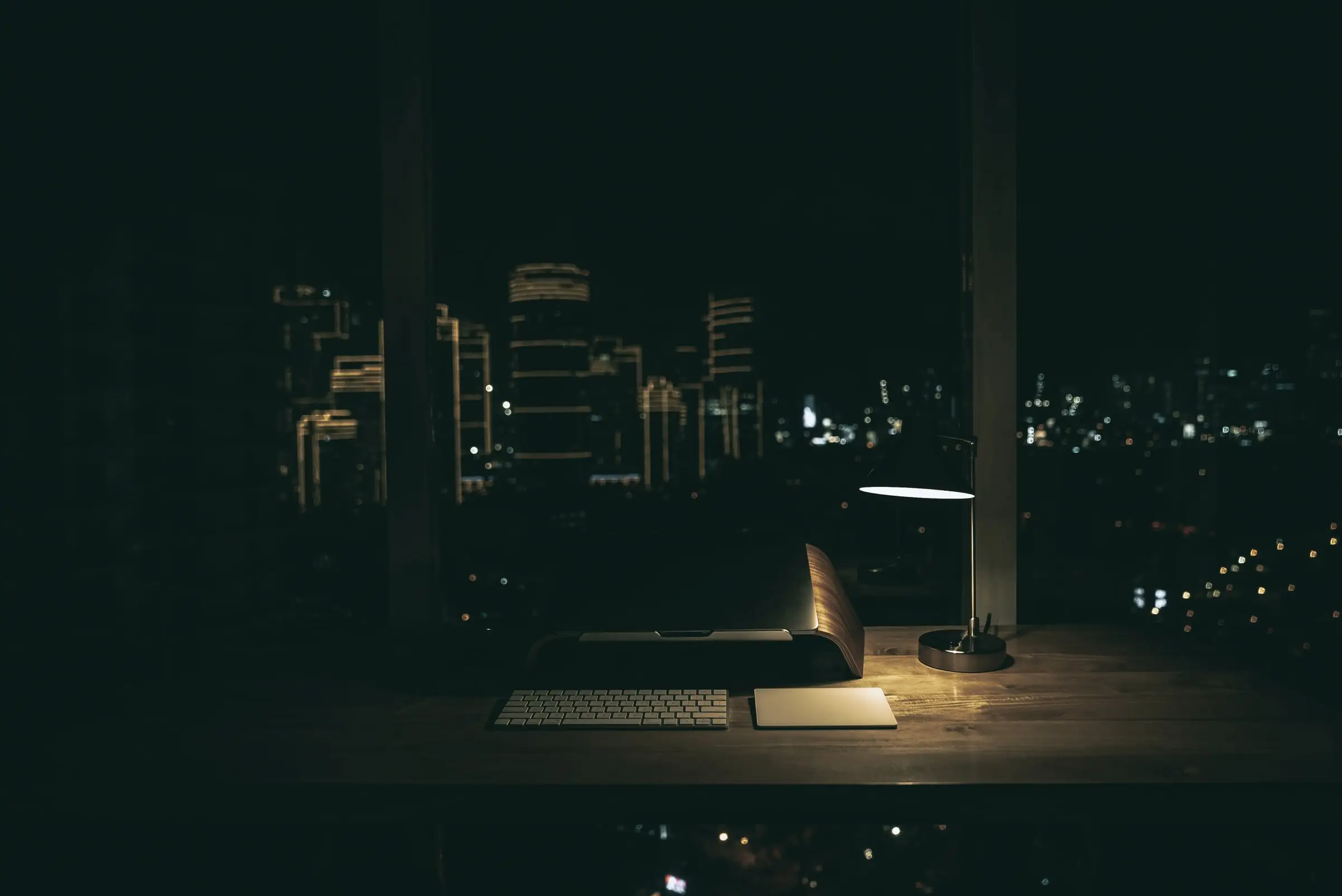 A desk lamp lights a keyboard on a desk by a window over a city at night
